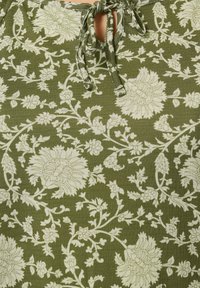 Zielona bluzka z tkaniny w kwiatowy wzór w kolorze kremowym. Posiada detal w postaci kokardy przy dekolcie. Teksturowana powierzchnia z misternymi wzorami kwiatów i liści.