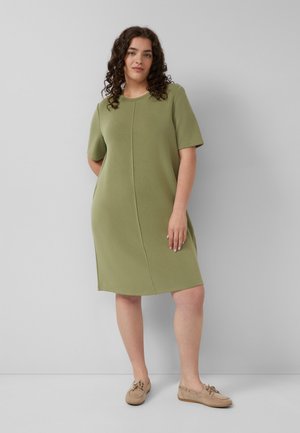 Kobieta o kręconych włosach, ubrana w oliwkowo-zieloną sukienkę z krótkim rękawem do kolan i beżowe mokasyny, stojąca na tle gładkiej szarej ściany.