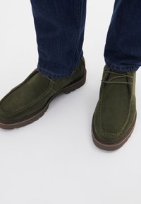 Mocasini verzi din suede cu un design de tip mocasin, prezentând detalii de cusătură și o talpă groasă maro, purtați cu blugi de culoare albastră închisă.