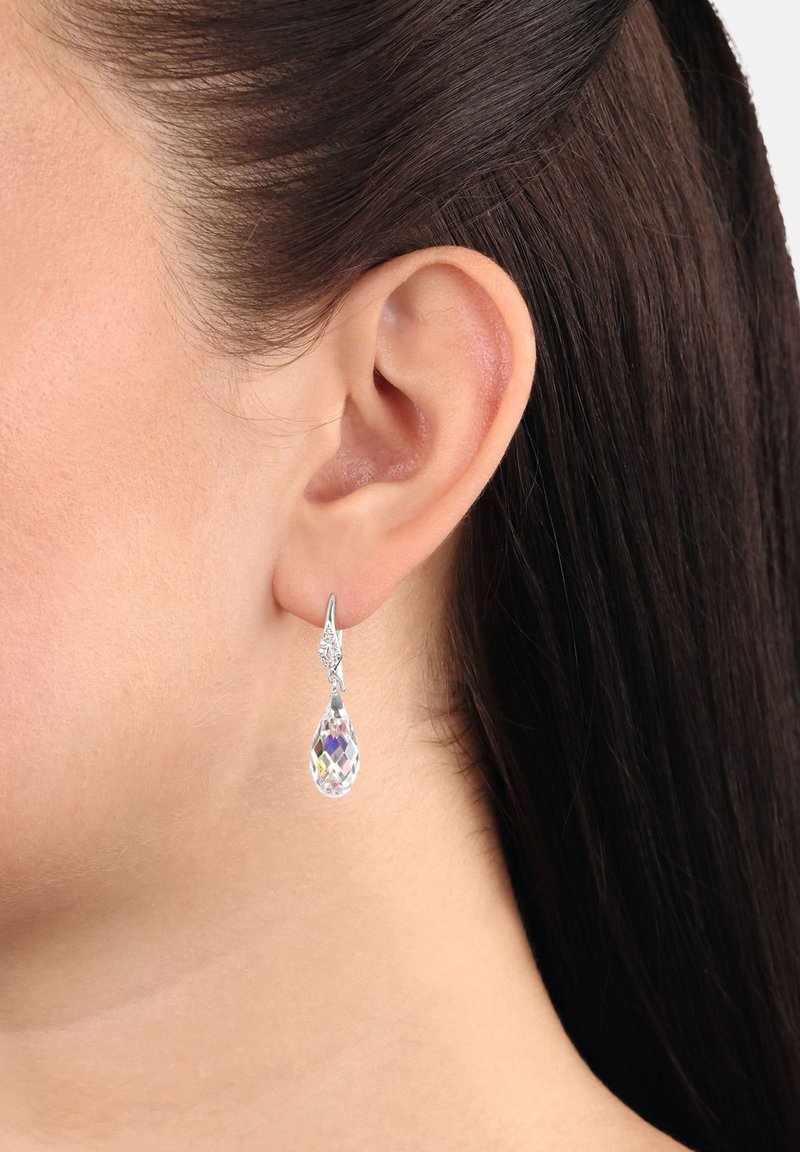 La oreja izquierda de una mujer lleva un pendiente de gota de plata con un colgante de cristal en forma de lágrima facetado y transparente.