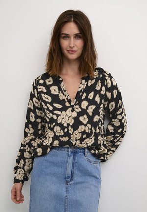 Sort bluse med beige blomsterdesign, V-hals og lange ærmer, kombineret med en lys blå denimnederdel. Teksturen ser glat ud.
