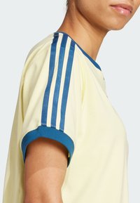 Svetlo žlté tričko s modrým rebrovaným golierom a akcentmi na rukávoch. Obsahuje tri modré vertikálne pruhy na každom pleci. Mäkká tkanina.