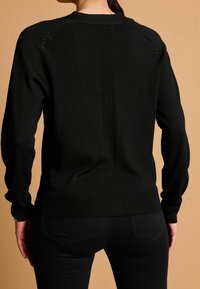 Sort strikket sweater med ribbede detaljer ved halsudskæringen og ryggen, med et struktureret mønster og tætsiddende ærmer, båret over sorte bukser.