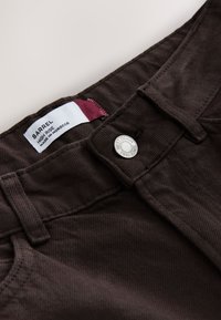 Close-up of brown high-rise denim jeans with silver button and visible label reading barrel high rise made in Morocco.