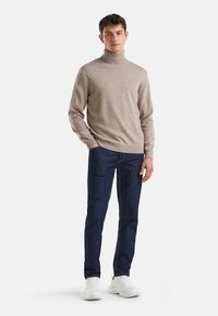 Maglione beige a collo alto in morbido tessuto a maglia, abbinato a jeans blu scuro e sneakers sportive bianche, in piedi su uno sfondo semplice.