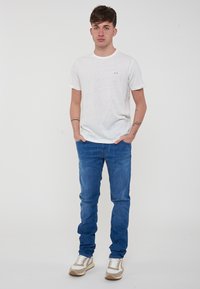 T-shirt bianco a girocollo con logo discreto, abbinato a jeans in denim blu. Il modello indossa sneakers chiare. Outfit semplice e casual.