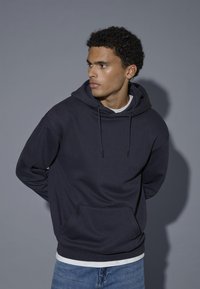 Ung mand med krøllet hår iført en mørk marineblå hættetrøje og blå jeans, der ser til sin venstre side mod en ensfarvet grå baggrund.