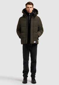 Tamsiai žalia pūkinė striukė su gaubtu, pačia su užtrauktuku priekyje, šoninėmis kišenėmis ir logotipo pleistru. Derinama su juodais džinsais ir auliniais batais.