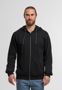 Zwarte zip-up hoodie gemaakt van zachte stof, met een trekkoordcapuchon, zijzakken en geribbelde boorden. Model draagt lichtblauwe broek.