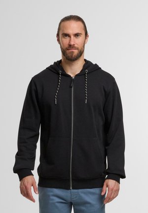Zwarte zip-up hoodie gemaakt van zachte stof, met een trekkoordcapuchon, zijzakken en geribbelde boorden. Model draagt lichtblauwe broek.