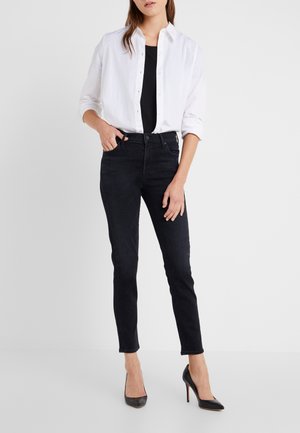 Femme portant un jean skinny noir taille haute, un top noir, une chemise blanche déboutonnée avec manches retroussées et des chaussures à talons noires.
