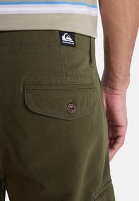 Close-up van olijfgroene shorts met een knopen achterzak en een Quiksilver-logo patch, gedragen met een beige gestreept shirt.
