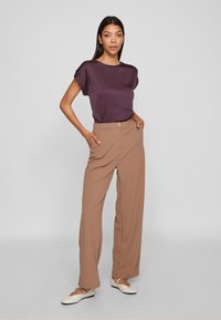 Γυναίκα που στέκεται με τα χέρια στις τσέπες, φορώντας μπεζ ψηλόμεσα παντελόνια καμπάνα, μωβ μπλούζα κοντομάνικη και άσπρα παπούτσια.