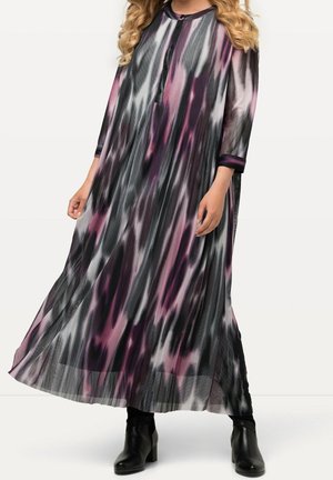 Longue robe plissée avec des motifs tie-dye noirs, blancs et violets, manches trois-quarts, encolure boutonnée, portée avec des bottines noires.