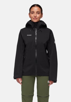 Frau trägt eine schwarze Kapuzenjacke mit Reißverschluss und olivgrüne Hosen, steht vor einem schlichten, hellen Hintergrund.