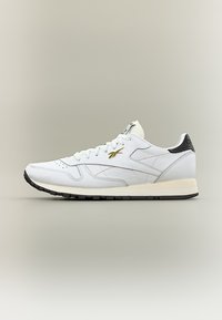 Λευκό δερμάτινο αθλητικό παπούτσι χαμηλού προφίλ με μαύρο τακούνι, χρυσό λογότυπο στο πλάι, λευκά κορδόνια και μαύρη λάστιχη σόλα.