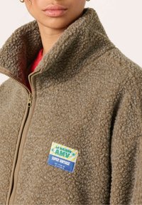 Ruskea fleece-zip-up-takki korkealla kauluksella, jossa on värikäs etiketti vasemmassa rinnassa ja pehmeä, tekstuuri. 