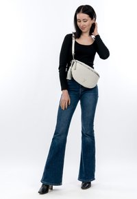 Naine, kes kannab musta pika varrukaga pluusi, laiade jalgadega siniseid teksapükse, musti saapaid ja valget ristiastmega kotti tussidega, seisab valge tausta ees.