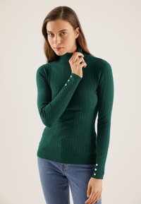 Green, knitted turtleneck sweater with ribbed texture, slim fit, and three decorative buttons on each cuff. Paired with blue jeans.