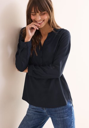 Cercasi camicia blu scuro con colletto alla coreana, maniche lunghe e spacchi laterali, abbinata a jeans in denim blu. Tessuto morbido e vestibilità rilassata.
