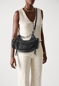 Black leather handbag with zippered pockets, silver hardware, and a chain strap. Features a curved shape and tassel accents.