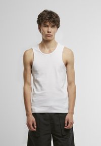 Weiße, ärmellose Tanktop aus Baumwolle, mit rundem Ausschnitt und einer einfachen, glatten Textur; getragen mit dunklen, locker sitzenden Shorts.