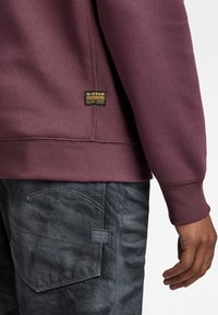 Bordowy sweatshirt z prążkowanymi mankietami, z naszywką G-Star Originals. W połączeniu z ciemnoszarymi jeansami, które mają subtelną fakturę i akcenty stebnowania.