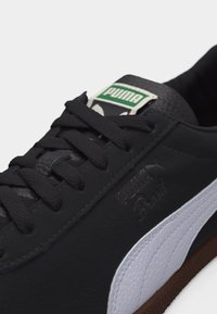 Κοντινό πλάνο μαύρου αθλητικού παπουτσιού Puma Brazil που δείχνει τα κορδόνια, την λευκή υφή στη πλαϊνή ρίγα, το ανάγλυφο λογότυπο και το πράσινο λογότυπο Puma στη γλώσσα.