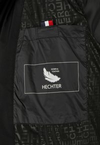 Melna jakas odere ar balto emblēmu, kurā attēlota spalva un teksts "DOWN & FEATHER HECHTER." Iekļauta sarkana, balta un zila auduma etiķete.