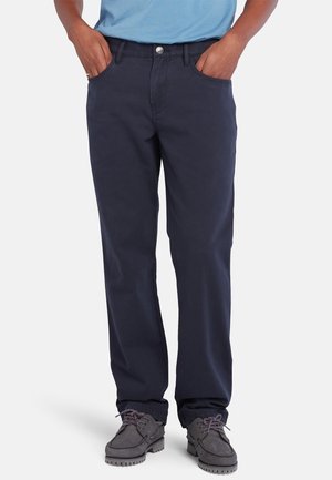 Mężczyzna w ciemnoniebieskich, prostych spodniach, szarych sznurowanych butach i jasnoniebieskiej koszuli, z rękami w kieszeniach.