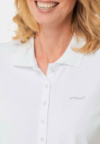 Weißes Poloshirt mit strukturiertem Finish, ausgestattet mit einer geknöpften Leiste und einem silberfarbenen bestickten Akzent auf der linken Brustseite.