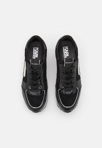 Zapatillas negras que presentan una mezcla de texturas mates y brillantes, con un diseño de cordones, puntera redondeada y detalles en blanco en el costado.