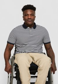 Striped polo shirt in navy and white with a collar, paired with beige trousers. Individual seated in a wheelchair, holding the armrest.