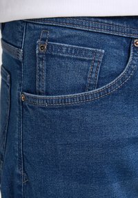 Jeans in denim di una tonalità blu media, con cuciture visibili, una piccola tasca per le monete e una texture liscia. In generale, un design aderente.