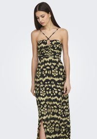 Mujer vistiendo un vestido maxi sin mangas, de color negro y verde oliva con un patrón, tirantes entrecruzados y una abertura frontal, de pie frente a un fondo liso.