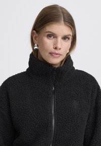 Mustumisega must fliistag teeb kõrge kraega, tekstuurse pinnaga ja väike logo rinnal. Omab esiotsas lukku ja vaba lõikega.