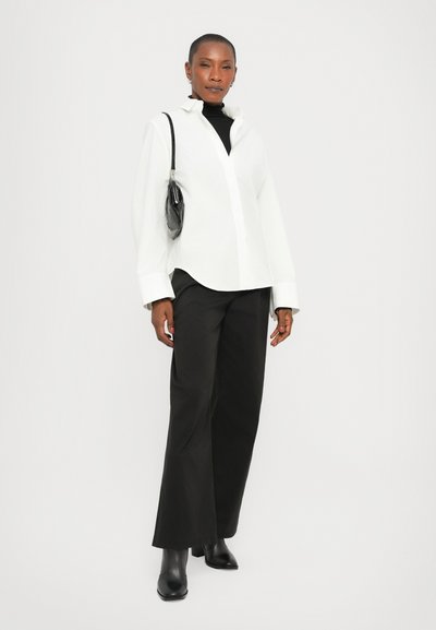 Chemise blanche à boutons avec manches longues, associée à un pantalon large noir et des bottines à talons. Accessoirisée avec un petit sac à main noir.