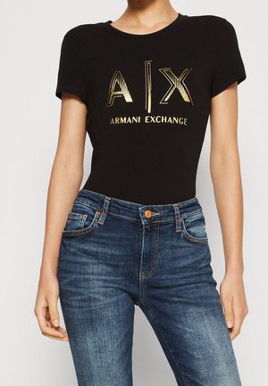 Kobieta w czarnej koszulce Armani Exchange z złotym logo, połączonej z niebieskimi dżinsami, pokazana od środka szyi do bioder.