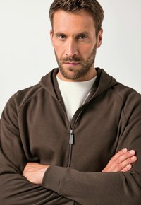 Felpa con zip marrone in tessuto texturizzato, dotata di cappuccio e dettaglio del logo sulla manica. Indossata sopra una maglietta chiara.