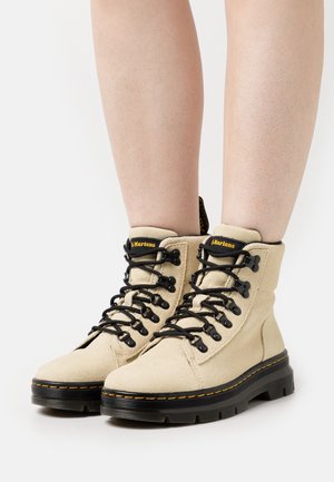 Béžové semišové kotníkové boty s černými hrubými podrážkami, černými tkaničkami a žlutým šitím, nošené na holých nohách na bílém pozadí.