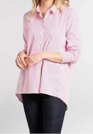 Kvinde iført en løs skjorte med rosa og hvide lodrette striber og opkappede ærmer samt mørke jeans, stående mod en ensfarvet baggrund.