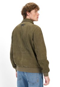 Oliven grøn suede bomberjakke med rib-strik ved håndled og kant, med en klapkrave og et mærke på nakken. Glat tekstur.