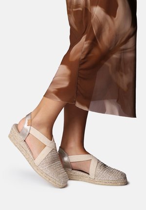 Füße in beigefarbenen, gewebten Espadrille-Sandalen mit elastischen Riemen und metallischen Fersenakzenten, kombiniert mit einem fließenden braunen Rock.