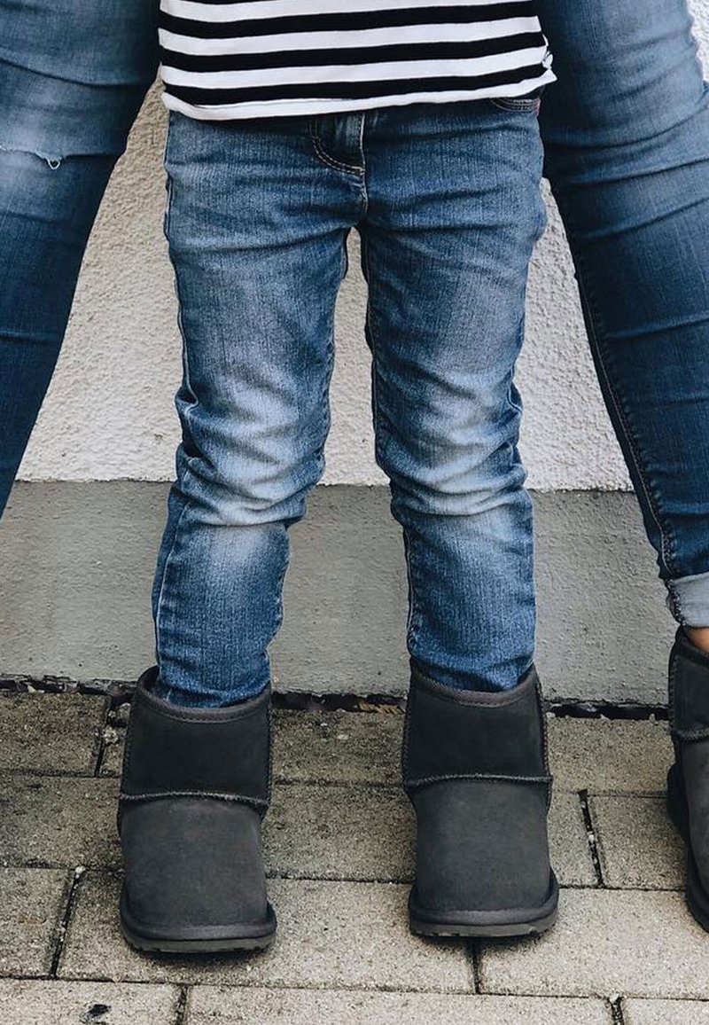 Dziecko w wyblakłych niebieskich skinny jeansach i szarych botkach za kostkę stoi obok osoby dorosłej, obie na kamiennej nawierzchni. Tło jest neutralne.