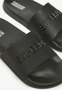 Zwarte slidesandalen gemaakt van textuur synthetisch materiaal met een brede band die voorzien is van reliëfletters "SUPERDRY". Plat zoolontwerp.