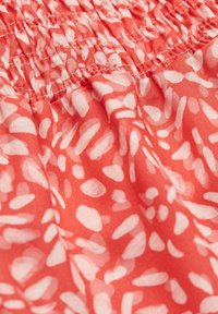 Gros plan sur un tissu corail avec des taches abstraites blanches irrégulières et un détail de couture élastique froncée.