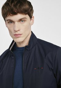 Granatowa kurtka zapinana na zamek z wysokim kołnierzem, z kontrastowym prążkowanym dekoltem i subtelnym haftowanym logo w czerwieni i czerni na klatce piersiowej.