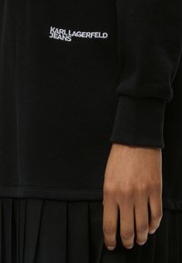 Μαύρο μακρυμάνικο τοπ με λευκή επιγραφή "KARL LAGERFELD JEANS", φορεμένο από άτομο με ορατό χέρι και μαύρη πλισέ φούστα.
