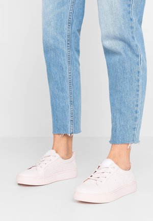 Person iført lyseblå korte jeans og lyserøde lave sneakers, stående på en lysegrå gulv mod en ensfarvet baggrund.