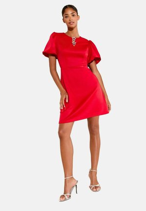 Rotes kurzes Kleid mit Puffärmeln, tailliertem Schnitt, glattem Stoff und einem sternförmigen Halsbesatz. Kombiniert mit silbernen Riemchensandalen.